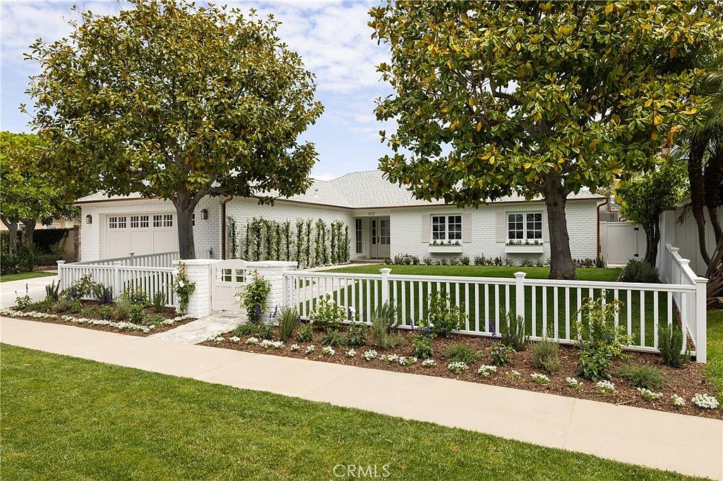 Front elevation of 1112 Pembroke Ln, a white single-story coastal house with a low gabled roof, a two-car garage on the left, fruit trees framing the entry, and a white picket fence holding a row of olive shrubs at the curb