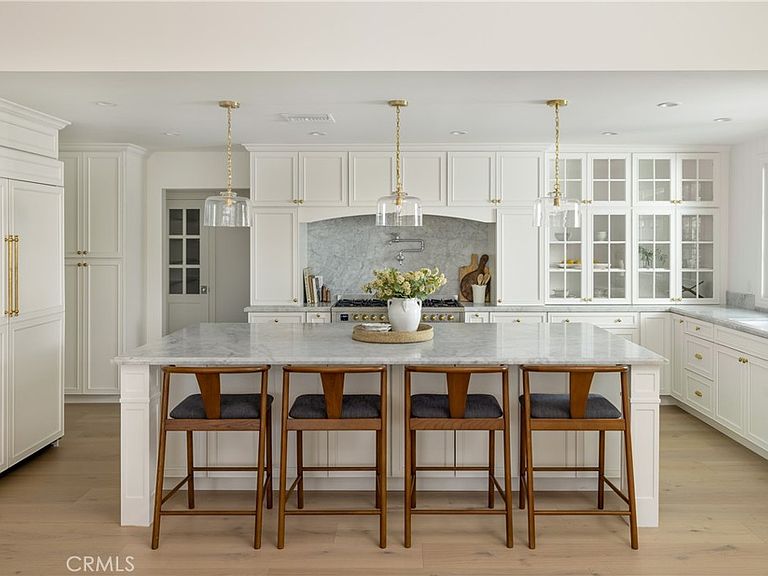Kitchen at 1112 Pembroke Ln with a long Bianco Carrara marble island, four wooden counter stools, three brass-and-glass pendant lights overhead, white inset cabinetry, and a marble-arched cooking alcove behind the range