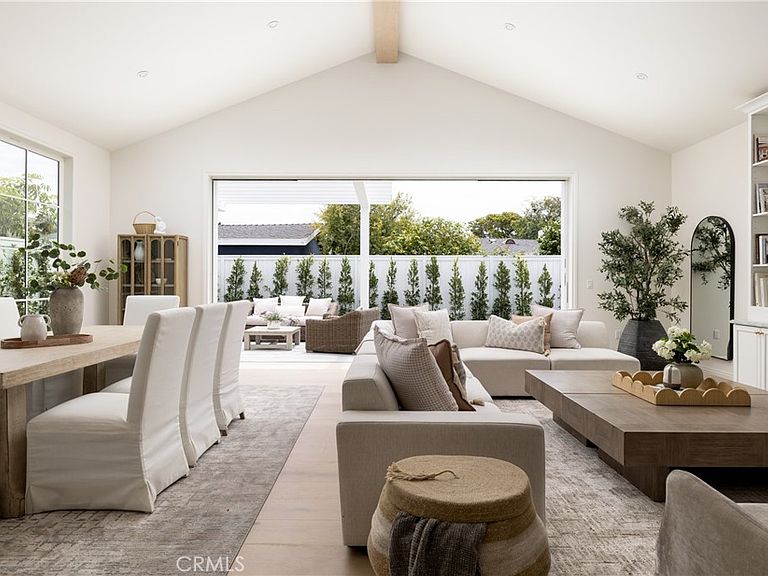 Living and dining area at 1112 Pembroke Ln with two vaulted ceilings, slipcovered armchairs at the dining table, a tall arched mirror to the right, and a wide window framing clipped boxwood hedges and a row of cypresses outside