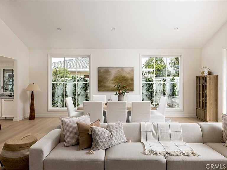 Living room at 1112 Pembroke Ln seen from behind a low-back sofa, looking through tall double-hung windows at a landscape painting and a dining table flanked by white slipcovered chairs and a tall wood cabinet