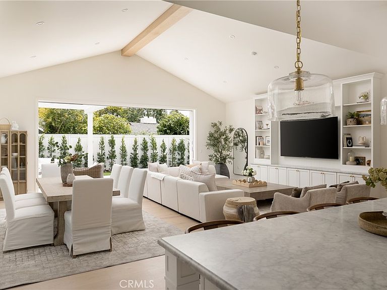Great room at 1112 Pembroke Ln with a sixteen-foot vaulted ceiling, a single exposed beam, and an eighteen-foot pocketing glass door opening past a long sofa onto a clipped boxwood hedge and the backyard beyond